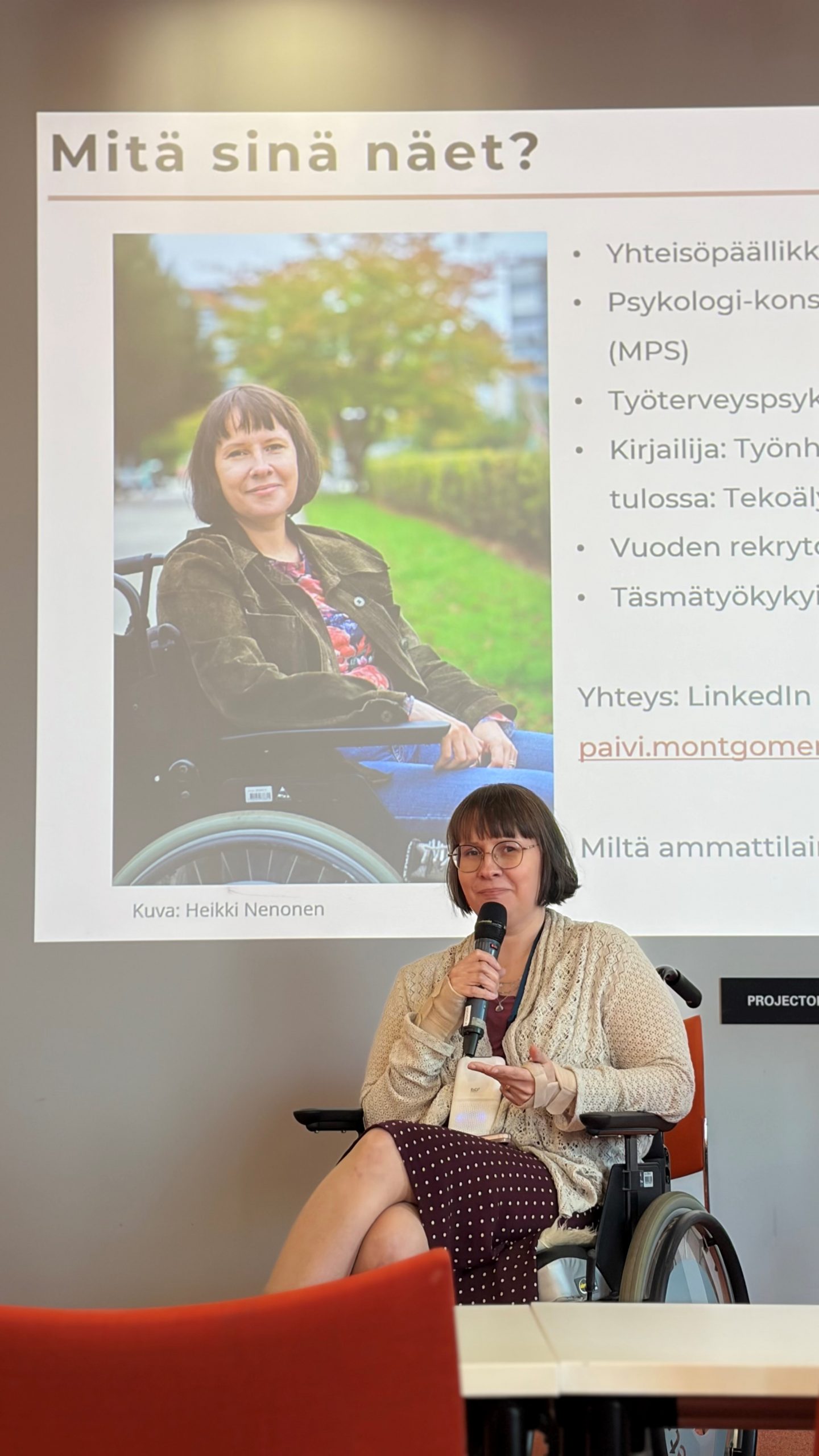 Henkilö puhuu yleisölle mikrofoni kädessä. Taustalla näkyy diaesitys.
